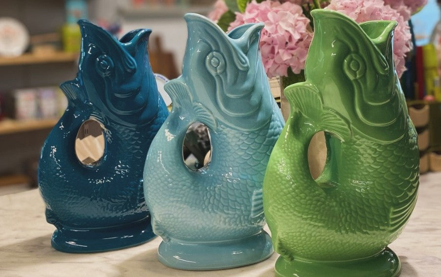 Three fish-shaped vases in blue, green, and teal on a table with flowers in the background.